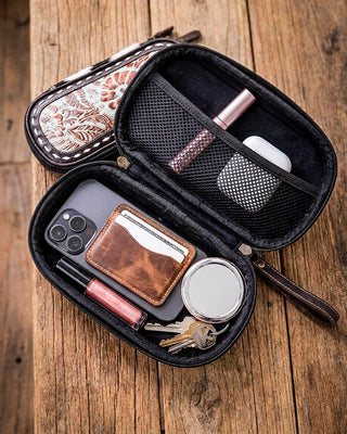An open black zippered phone wallet organizer case displayed on a rustic wooden table, showing its interior compartments filled with a phone, card holder, keys, lip gloss, and earbuds case. A second closed view of the case with a floral patterned exterior is shown behind it.