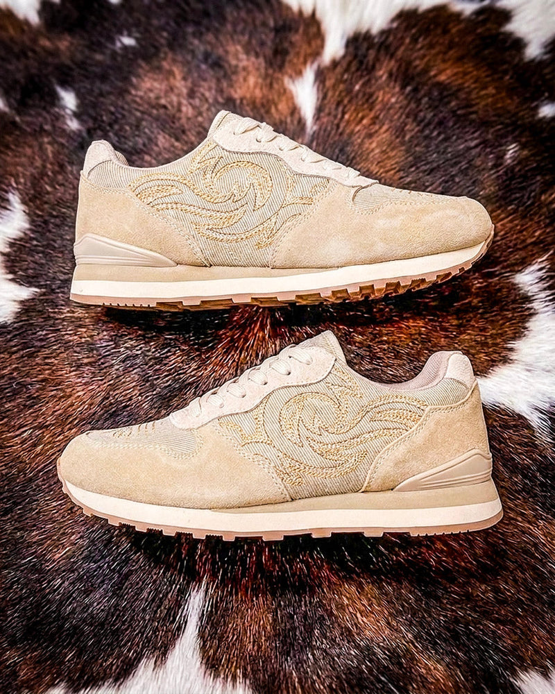 A pair of beige suede sneakers with tonal embroidered swirl patterns, displayed on a brown and white cowhide rug.