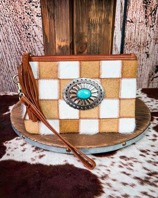 A western-style wristlet clutch bag with a brown and white checkered pattern made from different textures of hide and faux fur, featuring a large silver concho with a turquoise stone on the front, a brown leather wristlet strap, and a brown zipper closure. The bag is displayed on a round wooden platform with a rustic wood and cowhide background.