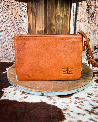 A tan leather wristlet clutch with a matching leather strap and gold-tone hardware, displayed on a round wooden platform with a cowhide rug and wood panel backdrop. The clutch features an embossed logo reading 'JC Western Co.
