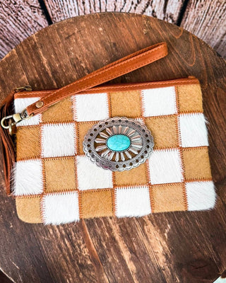 A checkered wristlet clutch featuring tan and white cowhide panels, a brown faux-leather wrist strap, and a large silver-toned concho with a turquoise center, displayed on a round wooden surface.
