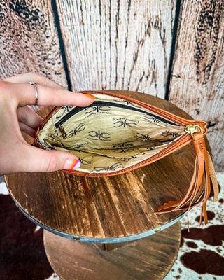 A hand holding open a tan leather zip pouch with a gold zipper and a printed interior, displayed on a rustic wooden stand against a weathered wood plank background.