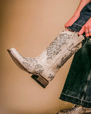 A person wearing a white western boot with intricate silver embroidery, holding the shaft of the boot while wearing dark blue jeans and a white long-sleeve shirt. Another foot wearing a matching western boot is visible at the bottom against a solid beige background.