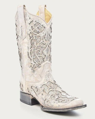 A single western cowboy boot in a distressed off-white leather, featuring intricate silver metallic embroidery and a dark gray stacked heel, shown against a light gray background.