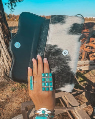 Hand holding a wallet with cowhide design, wearing turquoise jewelry, outdoors.