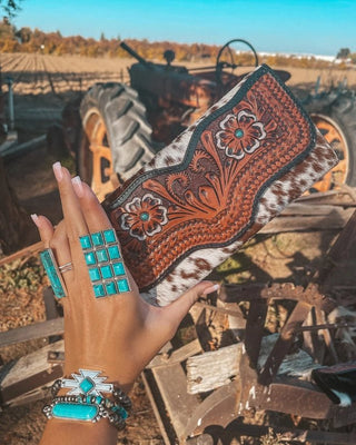 Hand holding a decorative clutch with intricate patterns and turquoise highlights, set against a rustic outdoor background.