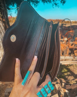 Hand holding a dark leather wallet with turquoise rings in front of a blurred outdoor background