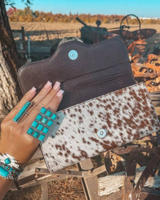 Hand holding a brown and white cowhide wallet against a rustic outdoor background.