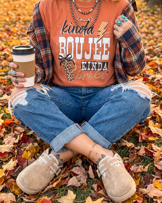 A person sitting cross-legged on a ground covered with autumn leaves, wearing a rust-orange graphic T-shirt layered over a plaid flannel shirt, ripped blue jeans, and beige slip-on shoes, while holding a takeaway coffee cup.