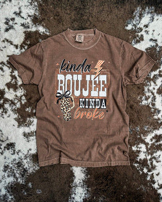 A brown graphic t-shirt laid flat on a brown and white cowhide rug. The shirt features a distressed graphic with the text 'kinda BOUJEE kinda broke' in white, black, and brown lettering, alongside a small leopard print and bow graphic.