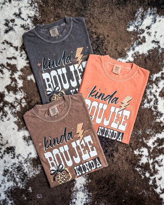 Three folded graphic T-shirts in charcoal gray, brown, and coral orange, arranged on a brown and white cowhide rug. Each shirt features the words 'kinda BOUJEE KINDA BROKE' in white and black with leopard-print and lightning-bolt graphics.