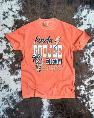A rust orange graphic t-shirt laid flat on a brown and white cowhide rug. The shirt features a graphic print that reads 'kinda BOUJEE kinda broke' in white, black, and beige fonts, accompanied by a small leopard print coffee cup illustration.