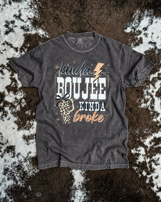 A pepper graphic t-shirt laid flat on a brown and white cowhide rug. The shirt features a graphic print that reads 'kinda BOUJEE kinda broke' in white, black, and beige fonts, accompanied by a small leopard print coffee cup illustration.