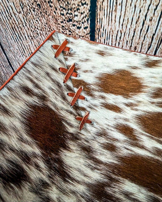A close-up of a cowhide bowl with brown and white mottled spots, finished with a reddish-brown stitched edge, laid on a rustic wood plank surface.