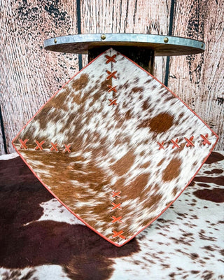 A square decorative tray with a cowhide pattern, featuring orange stitched details along the seams and a red-orange border, displayed on a rustic wood background.