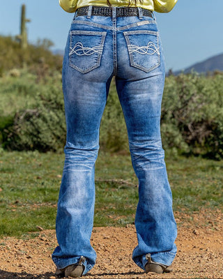 Person wearing blue jeans standing on a dirt path with greenery and mountains in the background