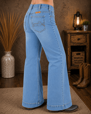 Light blue wide-leg denim jeans shown from the back on a model, standing in a rustic room with a wooden cabinet, boots, and a lantern.