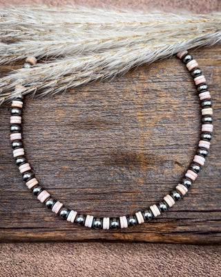 A beaded necklace with alternating black metallic beads, pale pink striped beads, and natural wood slice beads, displayed on a rustic wooden surface with dried grass.