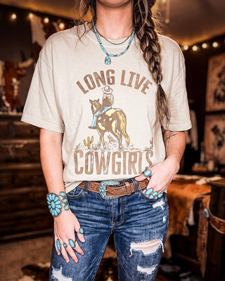 A woman wearing a beige graphic t-shirt with a brown and tan western print that reads 'LONG LIVE COWGIRLS' with an illustration of a cowboy on a horse. She is also wearing blue distressed jeans and turquoise jewelry in a rustic, warmly lit shop interior.