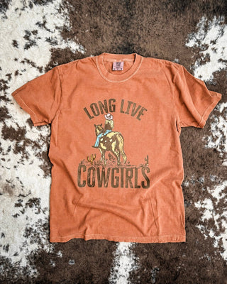 An orange short-sleeve graphic t-shirt laid flat on a brown and white cowhide rug. The shirt features a western-themed graphic with the text 'LONG LIVE COWGIRLS' and an illustration of a cowboy on a bucking horse.