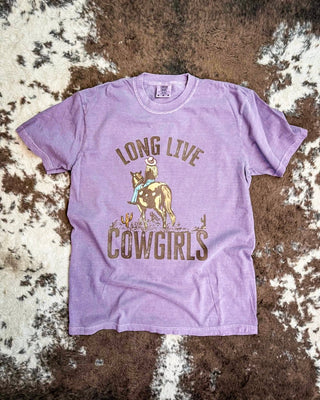 A lavender purple short-sleeve graphic t-shirt featuring a cowboy riding a horse with the text 'LONG LIVE COWGIRLS' printed on the front, laid flat on a brown and white cowhide rug.