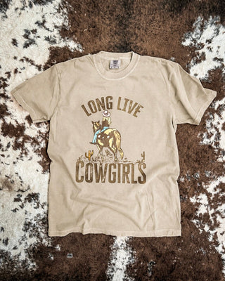 A beige graphic t-shirt featuring a cowboy riding a cowgirl with the text 'LONG LIVE COWGIRLS' printed on the front, laid flat on a brown and white cowhide rug.