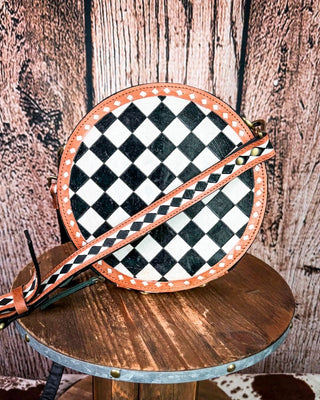 A round black and white checkered leather crossbody bag with orange-brown trim and a matching checkered strap, displayed on a rustic wooden stool against a wood plank wall.