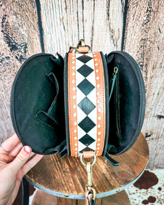 A black round zippered bag with a wide central strap featuring a black and white harlequin diamond pattern and orange side panels, held open by a hand over a rustic wooden stool.