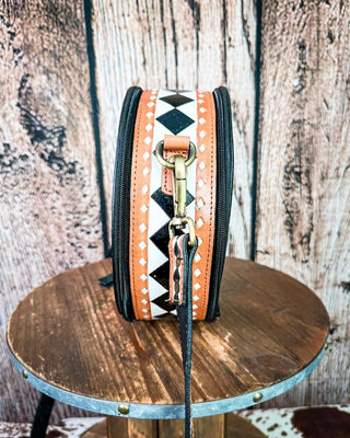 A small crossbody bag with a western-style checker pattern in orange, black, and white, displayed on a rustic wooden stool against a weathered wood plank background.