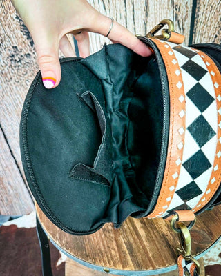 A close-up of a hand holding open a round black bag with a black-and-white harlequin patterned strap and tan leather trim, showing the black interior lining.