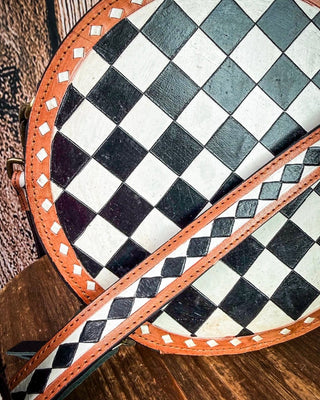 Close-up of a circular leather bag with a black and white checkerboard pattern, an orange-brown leather trim, and a matching checkerboard strap resting diagonally across the front.