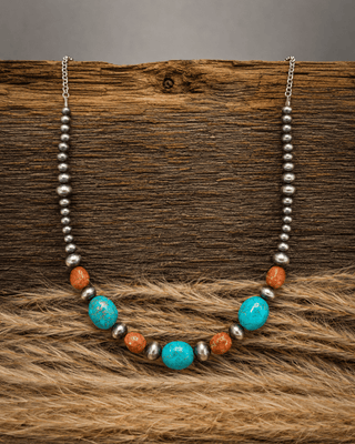 A beaded necklace with turquoise-blue oval beads, orange-brown round beads, and silver-toned spacer beads, displayed on a rustic wood surface with tan fur in the foreground.
