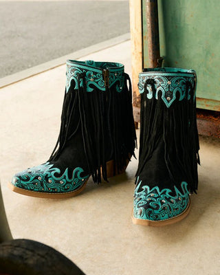 Black fringe boots with teal embroidery on a concrete floor.