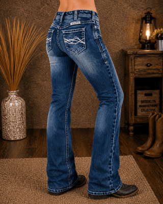A person wearing blue western flare jeans with decorative stitching on the back pockets, shown from the waist down in a rustic indoor setting with a wooden side table, boots, and a lamp.