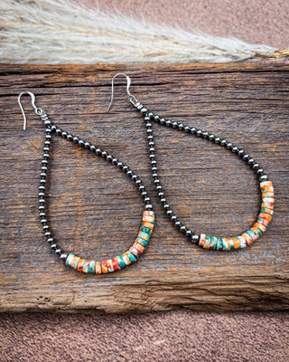 A pair of teardrop-shaped dangle earrings with black seed beads and a bottom section of colorful marbled beads in orange, teal, and cream, displayed on a rustic wooden surface.