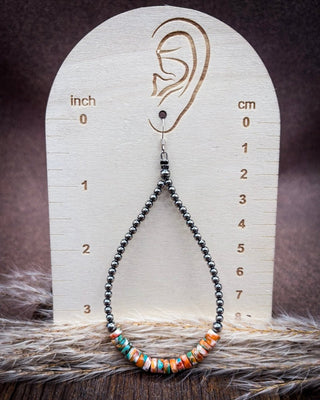 A beaded teardrop earring displayed on a wooden ear-measuring prop. The earring has dark gray beads leading to a cluster of colorful striped beads in orange, teal, white, and red.
