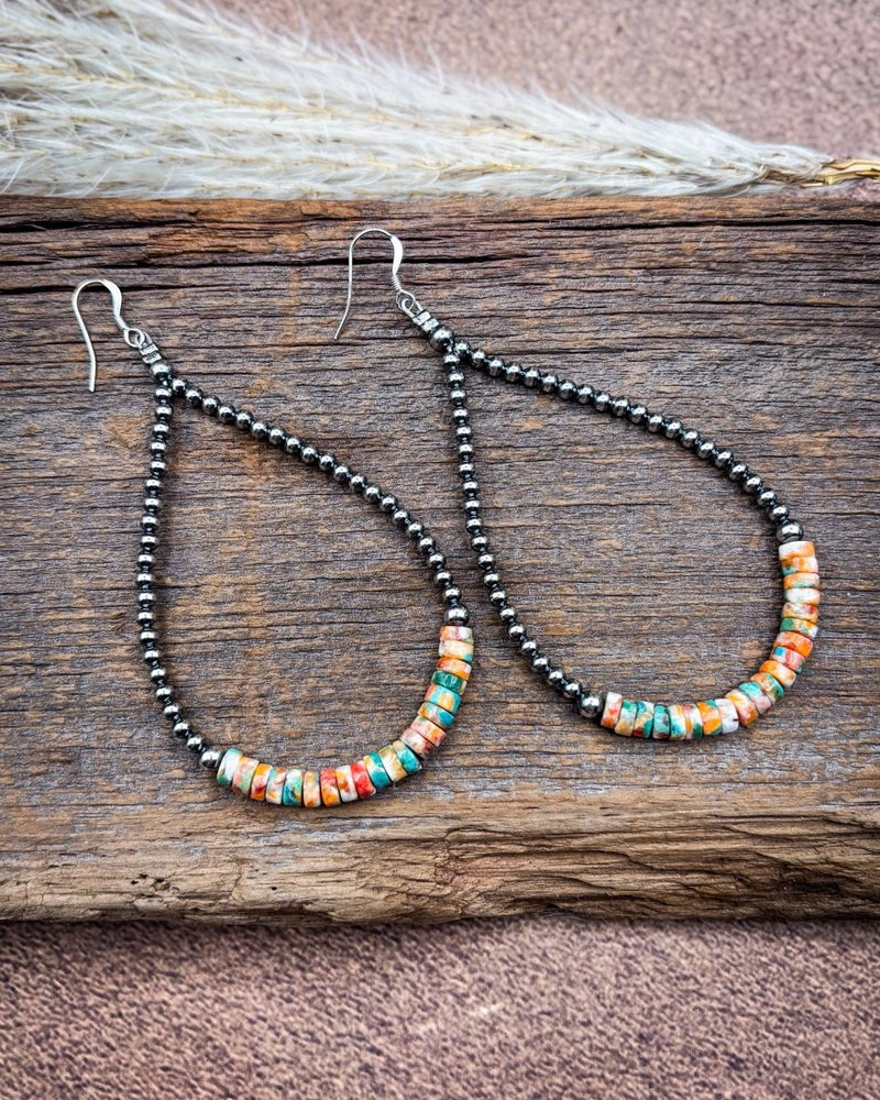 A pair of teardrop-shaped dangle earrings with black seed beads and a curved row of colorful stone-like beads in orange, turquoise, white, and red, displayed on a rustic wooden surface.