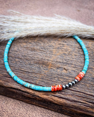 A beaded necklace with turquoise-blue beads, red-orange striped beads, and metallic silver beads, displayed on a rustic wooden surface with dried grass in the background.