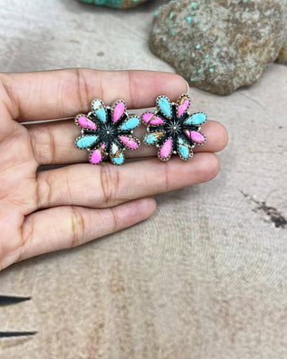 A pair of handmade flower-shaped stud earrings held on a person's fingers. The earrings feature turquoise blue, hot pink, and black enamel petals with silver-tone metal borders, set against a neutral textured background with rocks.