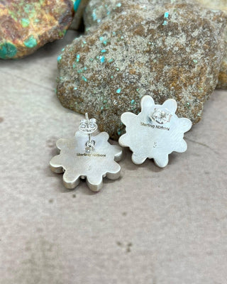 A pair of sterling silver flower-shaped stud earrings with a textured finish and a small clear crystal on the back, displayed on a neutral surface with decorative rocks in the background.