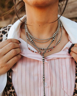 A close-up lifestyle shot of a person wearing a layered silver beaded necklace with a long drop pendant over a pink striped button-up shirt and a leopard print jacket.