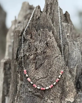 A silver chain necklace with a curved row of pink and white beads, draped over a textured piece of weathered wood.