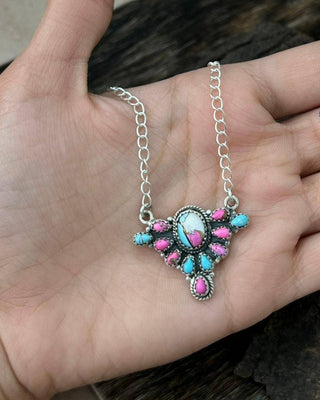 A silver-toned necklace with a Southwestern-style pendant featuring turquoise and pink stones, held in a person's hand.