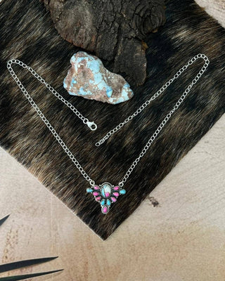 A silver chain necklace with a colorful pendant featuring pink, blue, and white details, displayed on a dark brown fur pad with a piece of wood and a turquoise stone.