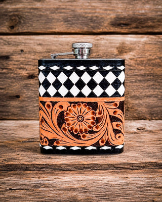 A rectangular metal flask with a silver screw cap, decorated with a black and white diamond pattern on the upper half and a carved orange floral scroll design on the lower half, set against a rustic wooden background.