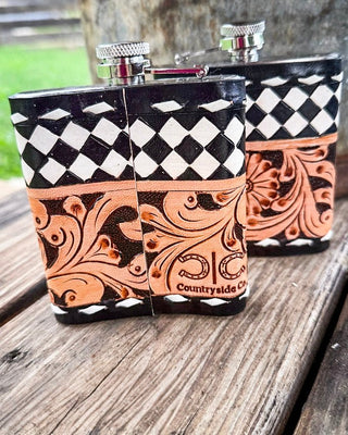 Two hand-tooled leather hip flasks with silver metal caps, featuring a black and white harlequin pattern on the top and a copper-orange tooled floral section on the bottom, displayed on a rustic wooden table.