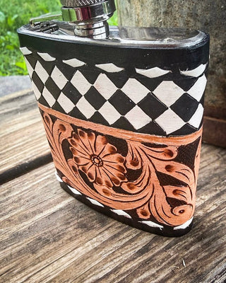 A small rectangular metal flask with a silver cap, decorated with a black and white harlequin pattern on the upper half and a carved copper-toned floral scroll design on the lower half, photographed on a rustic wooden surface outdoors.
