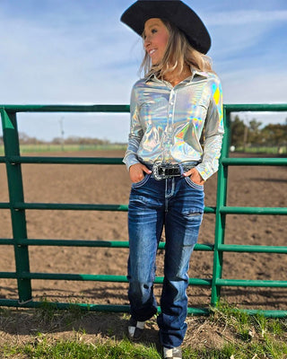 A woman wearing a shiny silver long-sleeve button-up shirt, blue jeans, a black cowboy hat, and a black belt stands outdoors in front of a green metal fence and dirt field.