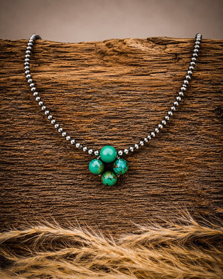 A beaded necklace with a cluster of green stone beads at the center, displayed on a textured piece of wood with dried grass in the foreground.