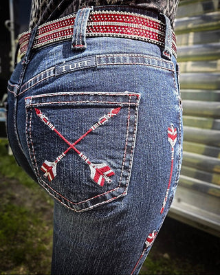 Close-up of the back of blue denim jeans worn by a person, featuring red and white embroidered crossed arrows on the back pocket and a matching red-and-white embroidered belt.